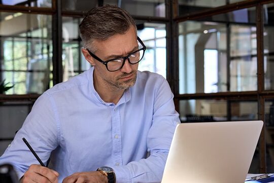 Ein Geschäftsmann sitzt im Büro am Laptop und nimmt an einem Web-Seminar teil, er macht sich Notizen.